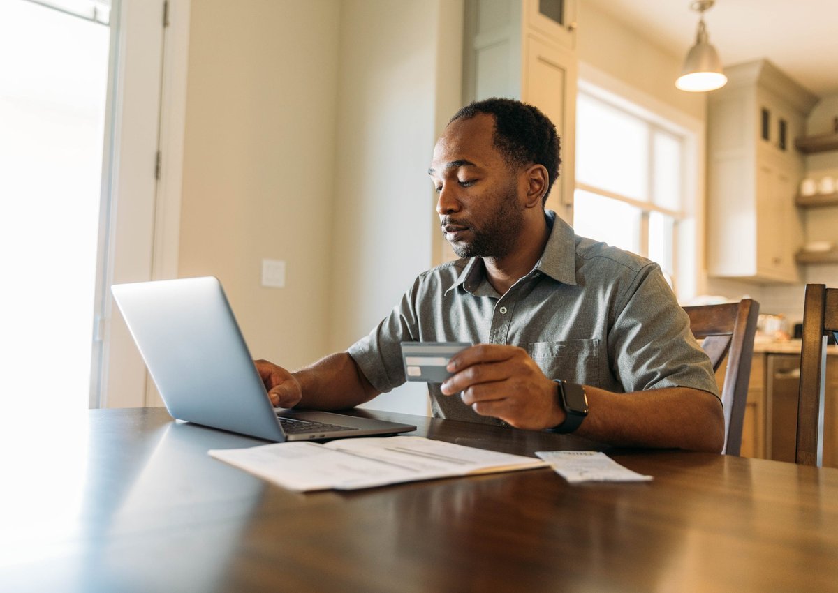 A man sits at a table, holding a credit card and paying his bills online.