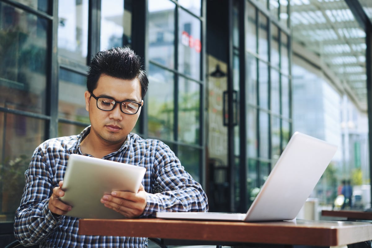 A man reads on a tablet with an open laptop on the table.