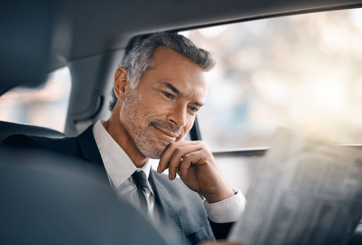 A man sits in a car, reading a newspaper.