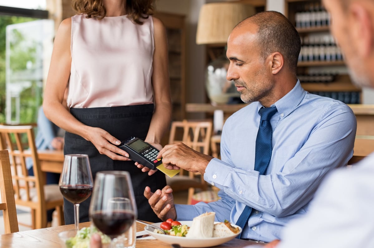 A businessman sits at a table in a restaurant and reads a credit card into a card reader held by a waitress.