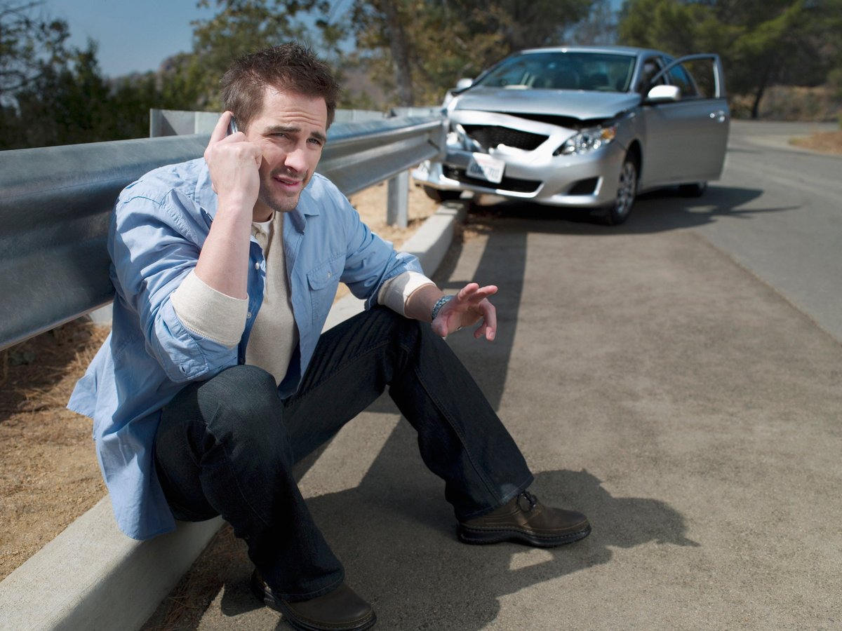 Man using his cellphone on the side of the road with a wrecked vehicle behind him.