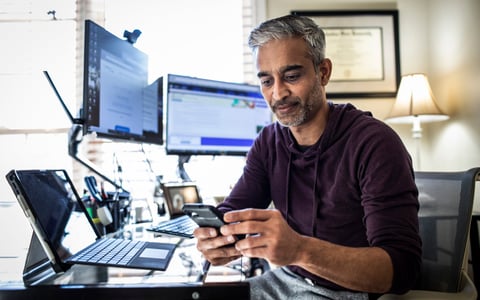 Man using is cell phone while sitting at desk with laptop and dual monitors.