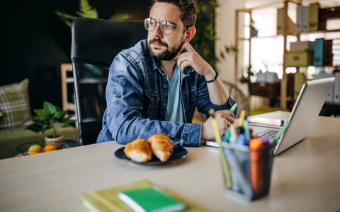 Man with glasses using laptop looks away appearing to be in thought.