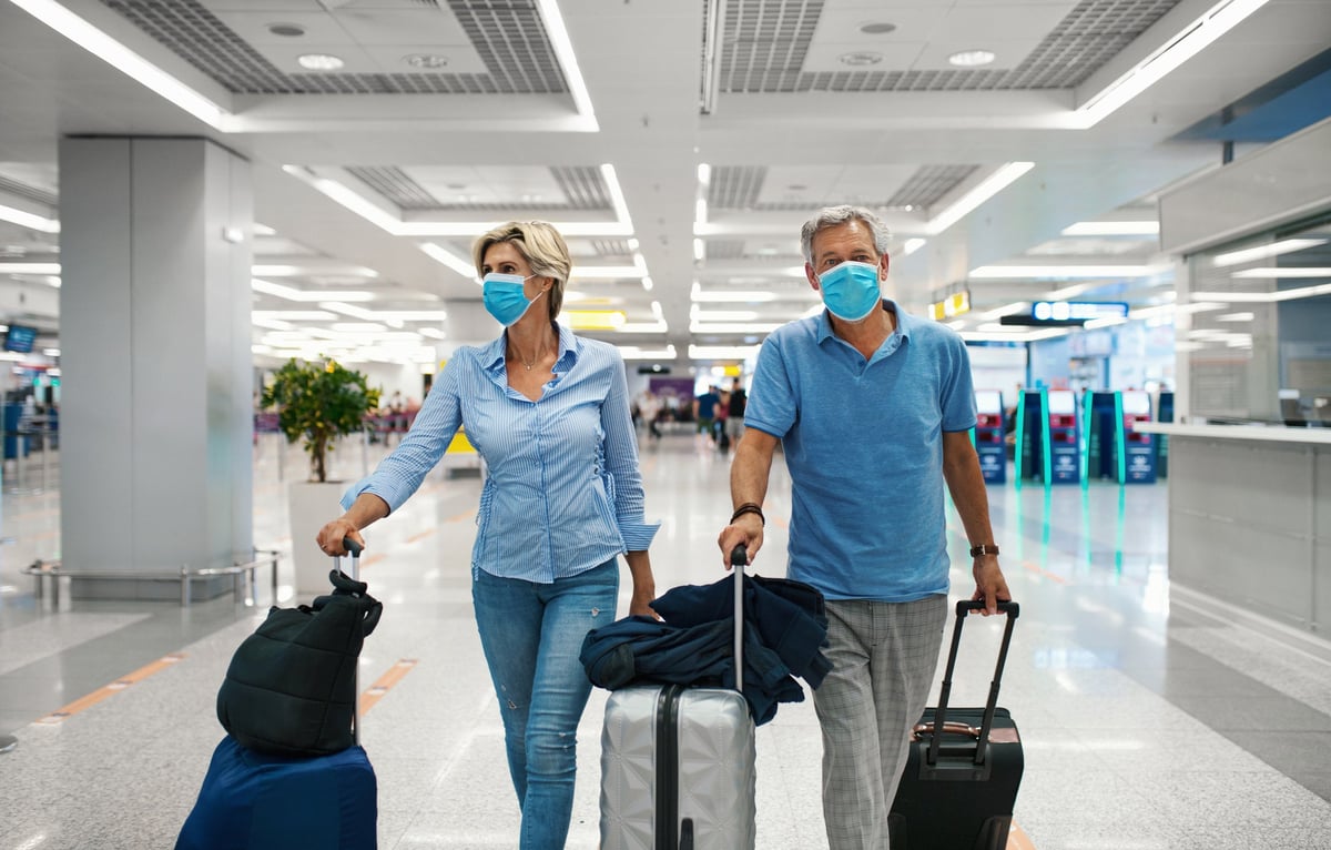 Mature couple walking through airport with luggage and masks.
