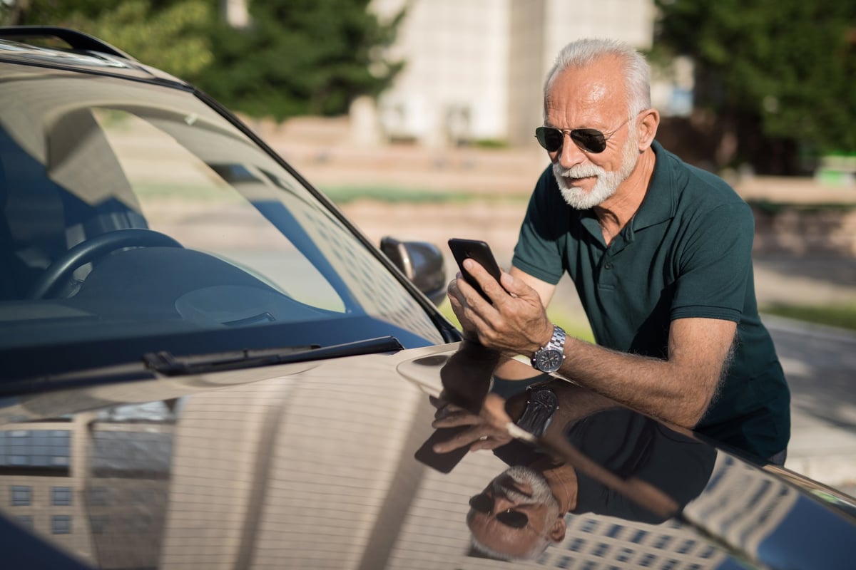 A mature man leans on his car as he looks at his phone.