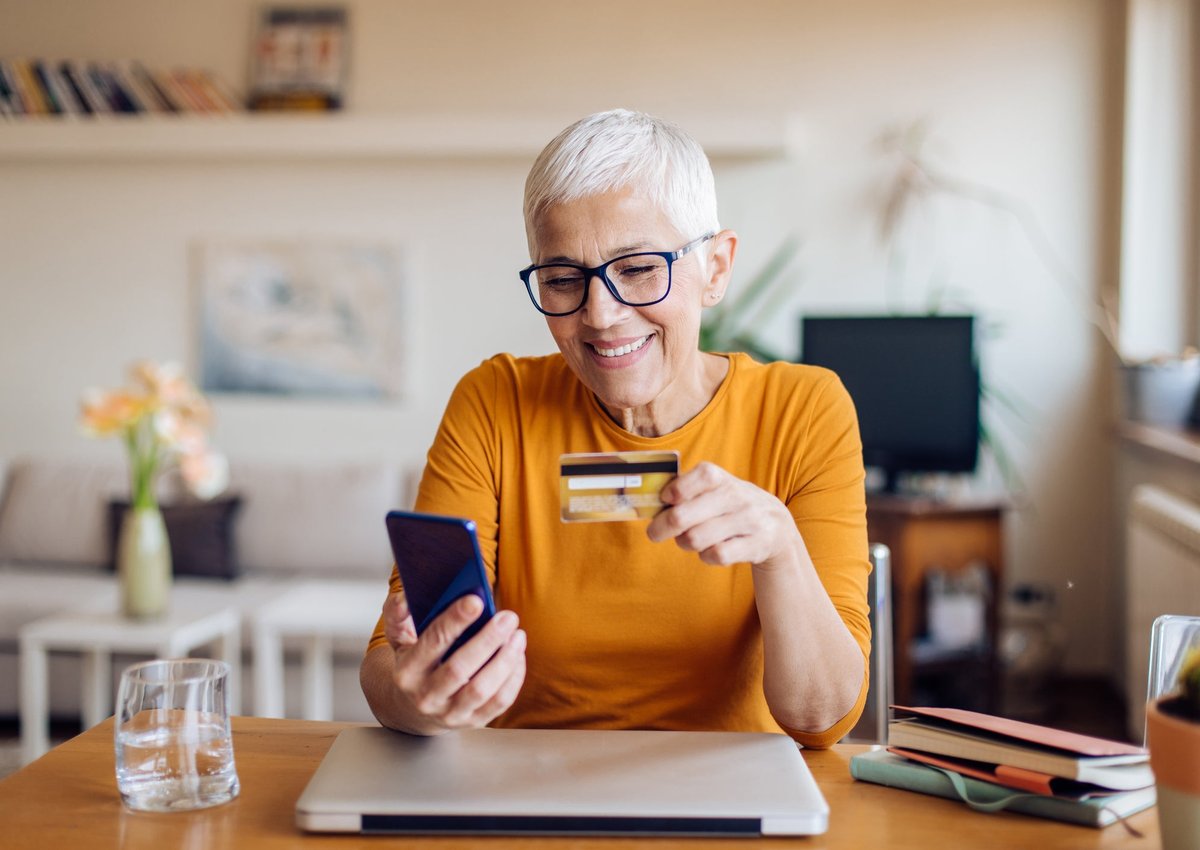 A mature woman smiles while sitting at her table. She is holding a cellphone and a credit card.