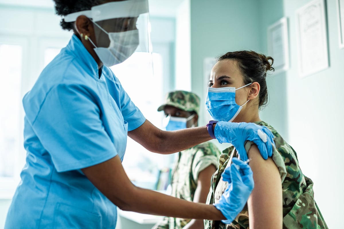 A medical staff member in a face shield provides an injection to a patient wearing a mask.