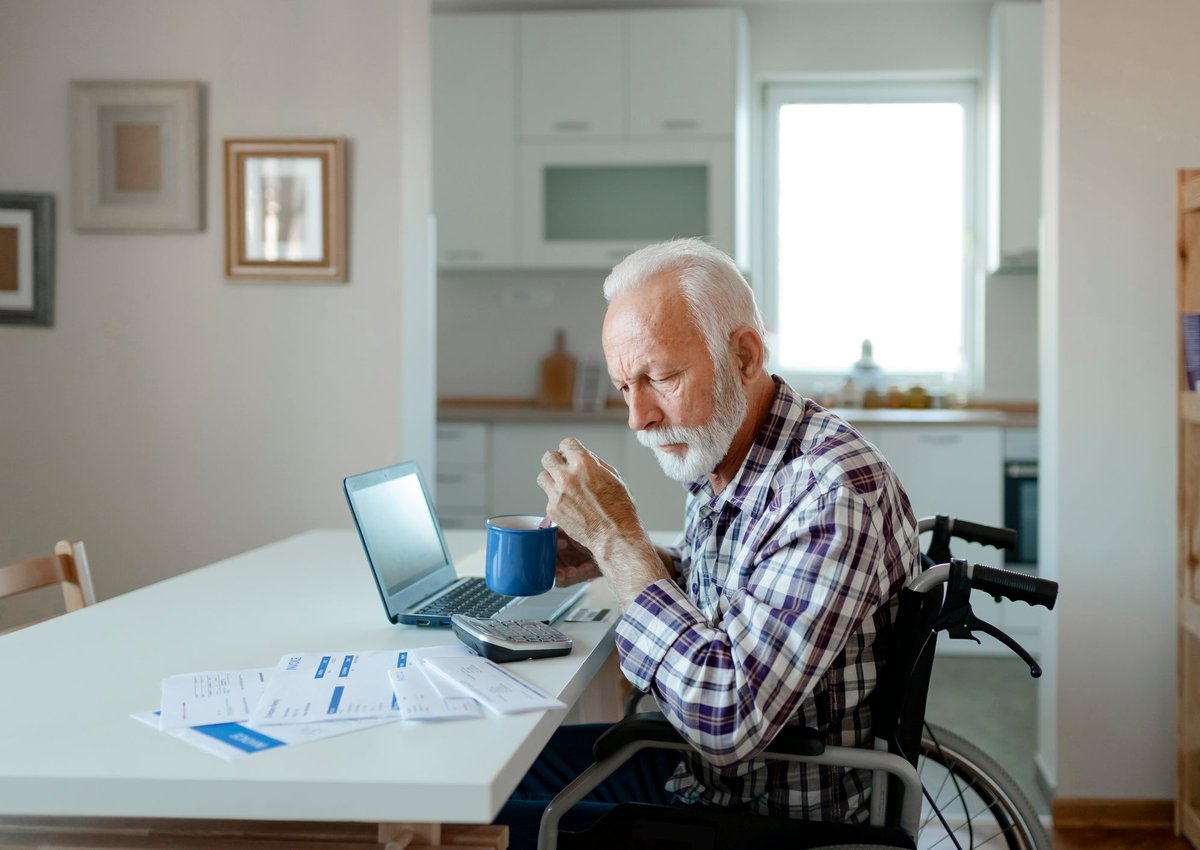 An older gentleman sits in a wheelchair at his kitchen table, looking over bills.