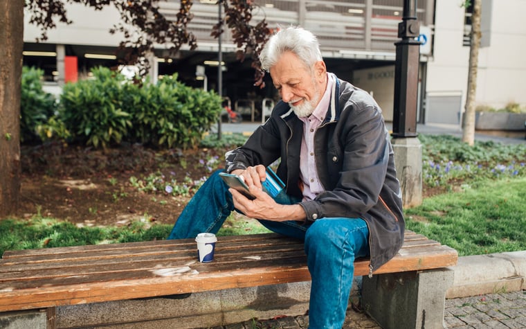 Un señor mayor se sienta en un banco del parque, mira su teléfono mientras sostiene su tarjeta de crédito.