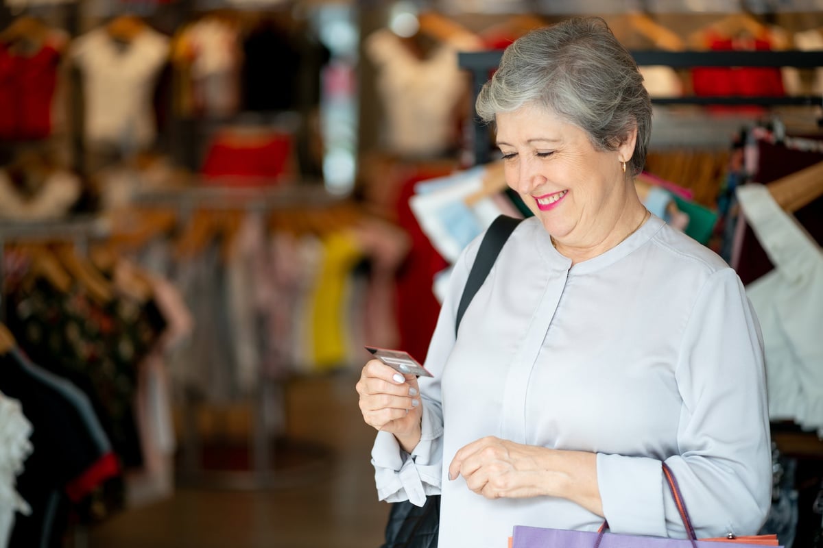 An older woman stands in a clothing store, looking happily at her rewards credit card.