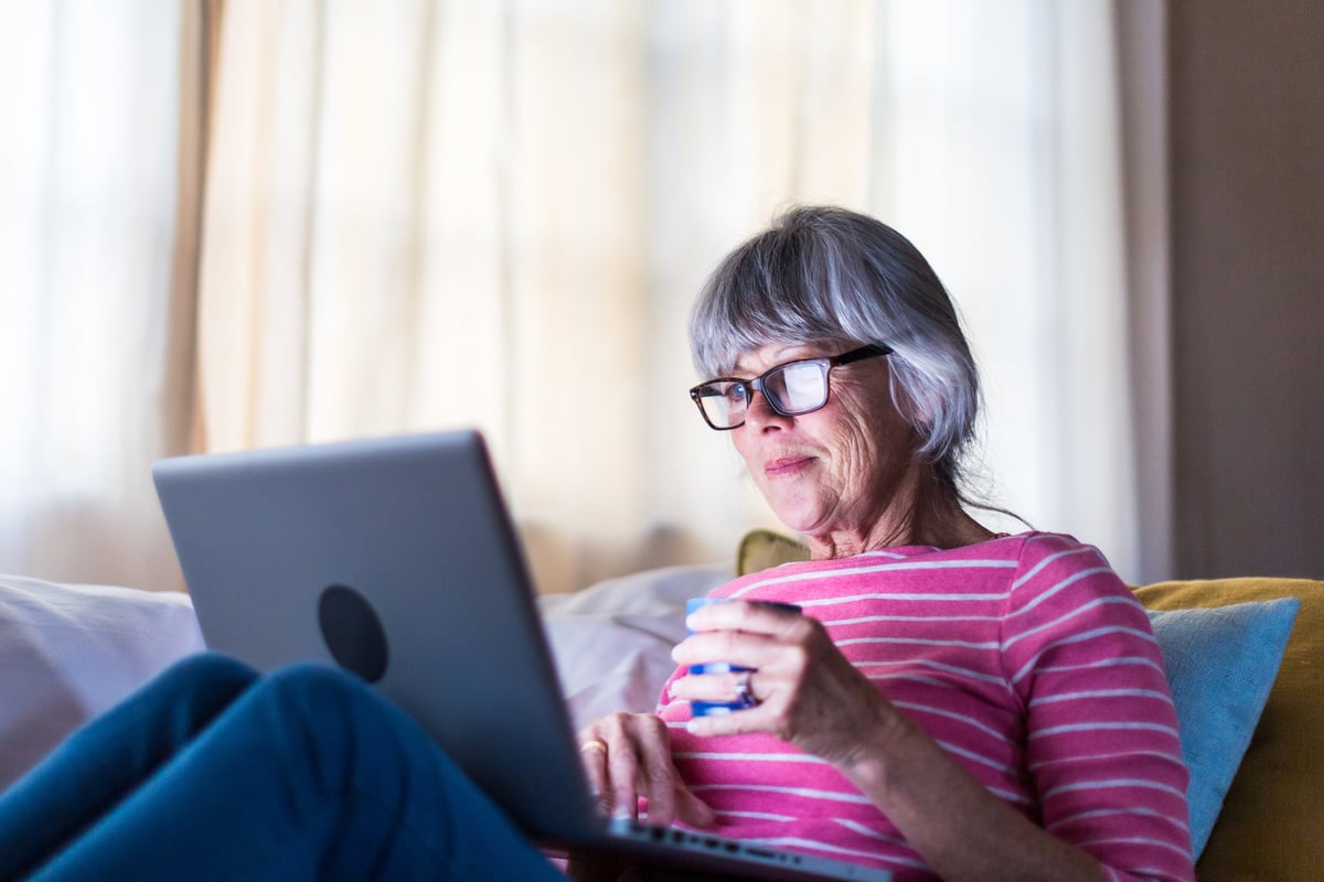 An older woman sits on her couch, looking at her laptop.