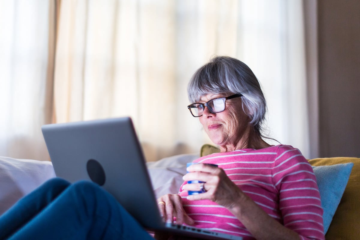 An older woman sits on her couch, looking at her laptop.