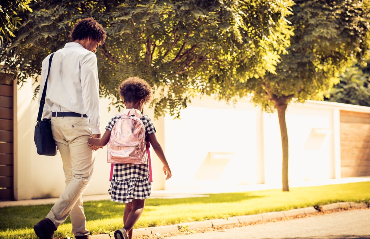 A father walking hand-in-hand with his young daughter wearing a backpack.