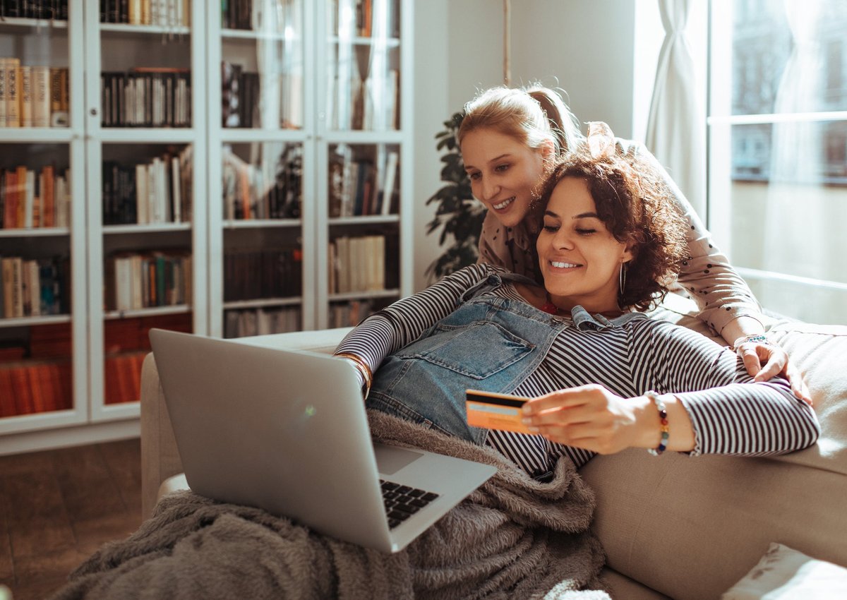 Pregnant woman holding credit card while using laptop with another woman looking over her shoulder.