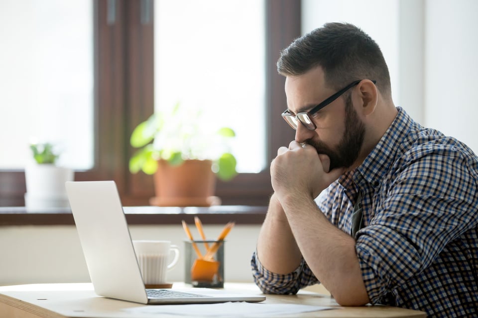 A man sitting at a desk with his laptop open and looking stressed with his chin in his hands.