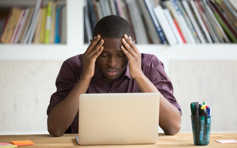 A person holds his head in his hands and looks frustrated at his laptop.