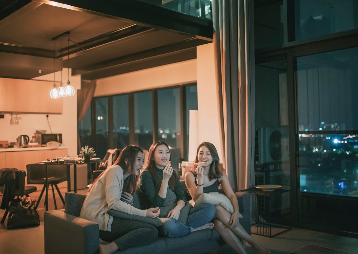 Three friends sit on a sofa in an apartment, sharing pizza.