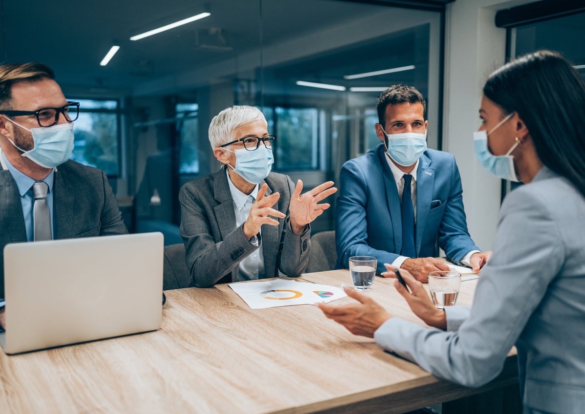 Three masked professionals interviewing masked woman in office setting.
