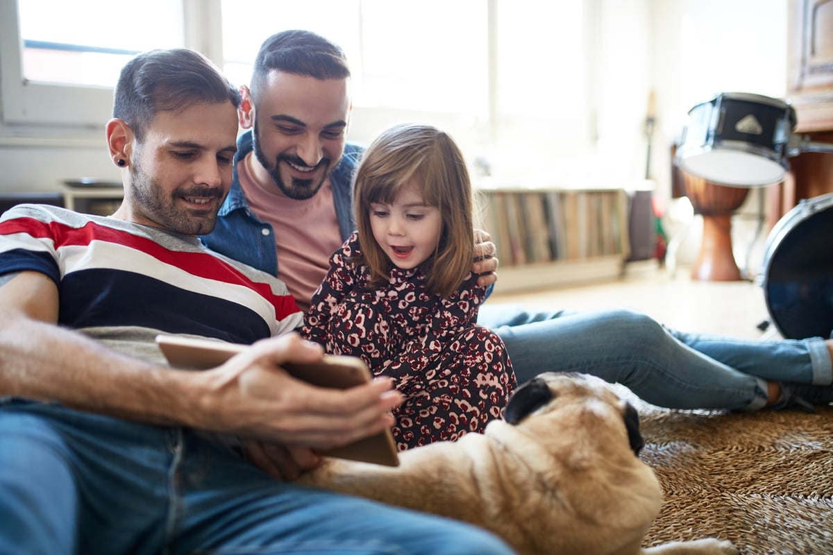 Two dads are on the floor with their daughter, looking at a tablet.