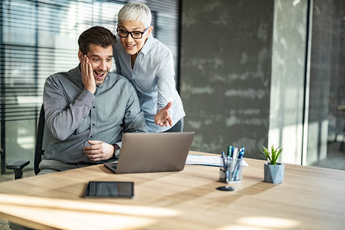 Two people in an office read the news on a computer and look pleasantly surprised.