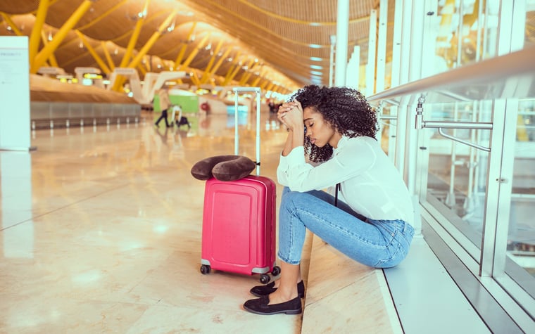 An upset woman at the airport sitting by a window next to her suitcase.