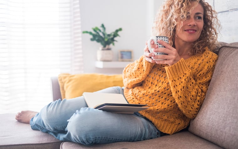 A woman sitting comfortably on her couch while drinking coffee and reading a book.