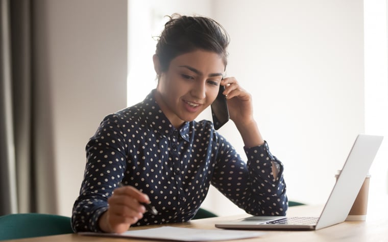 A young woman is talking on the phone with a laptop and documents.