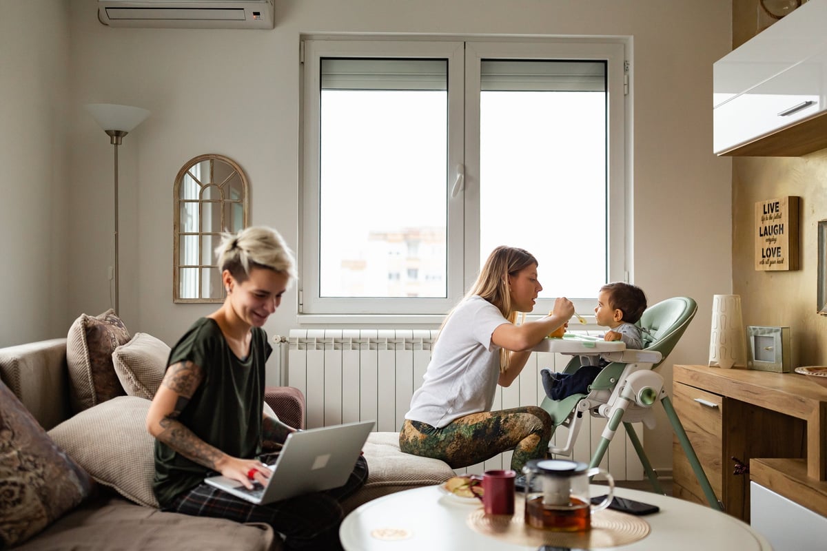 A family sits in their living room. While a woman works on her laptop, her wife feeds their baby.