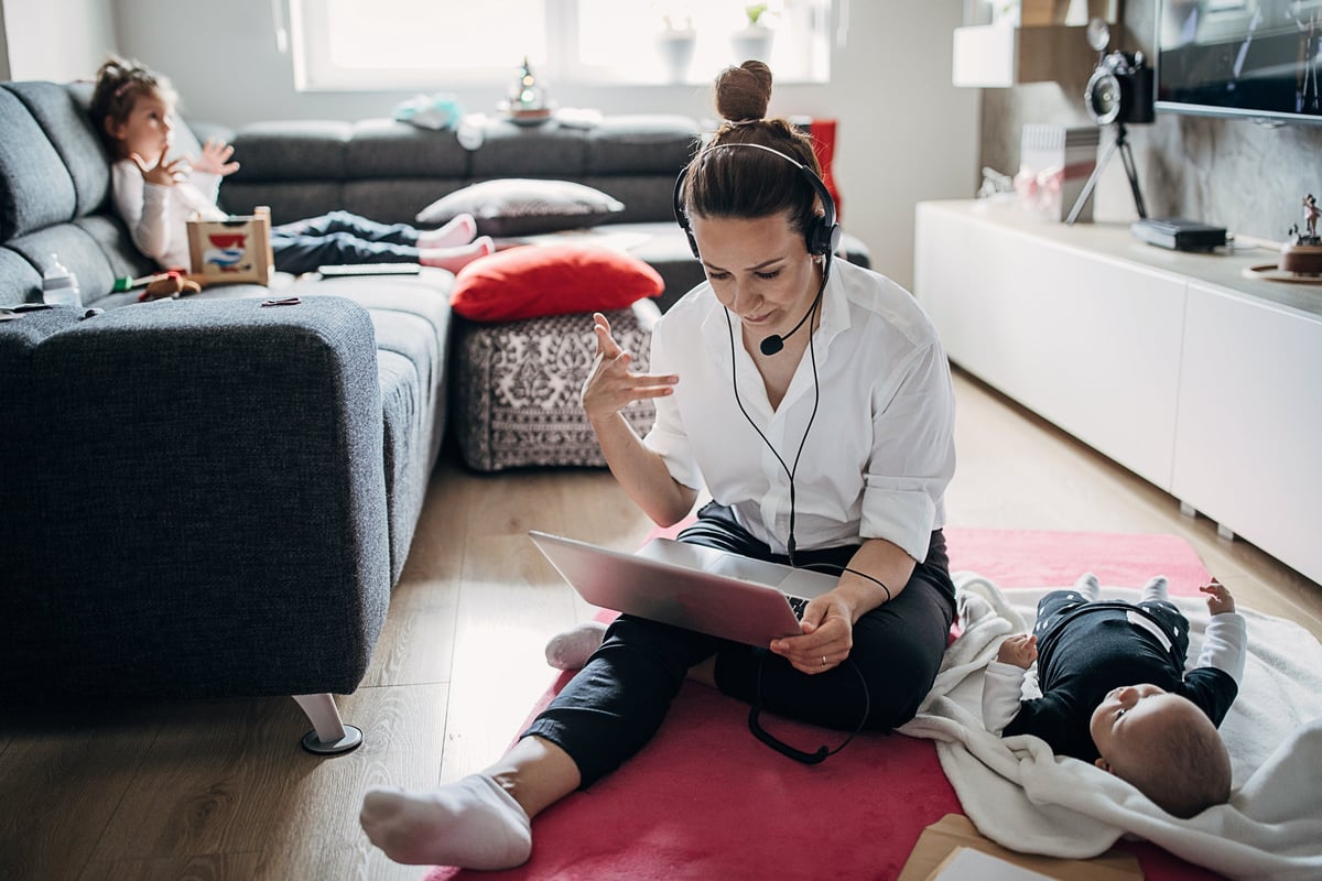 A working mom sits on the floor with her computer and infant daughter while her other daughter watches TV.