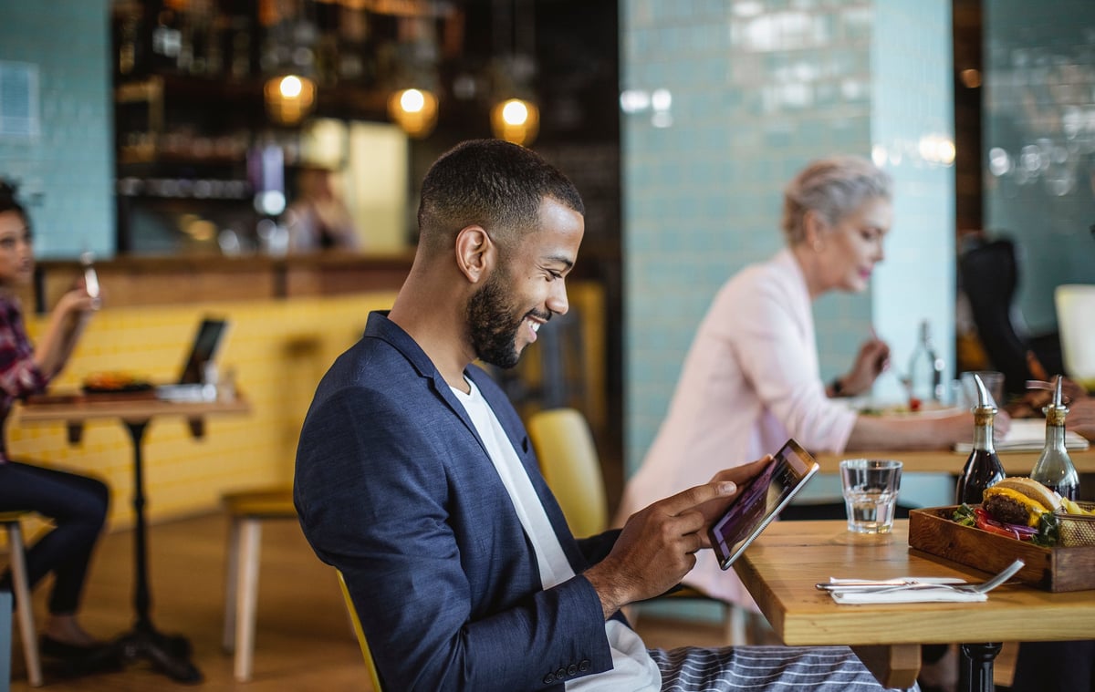 A young businessman sits in a coffee shop, reading on a tablet.