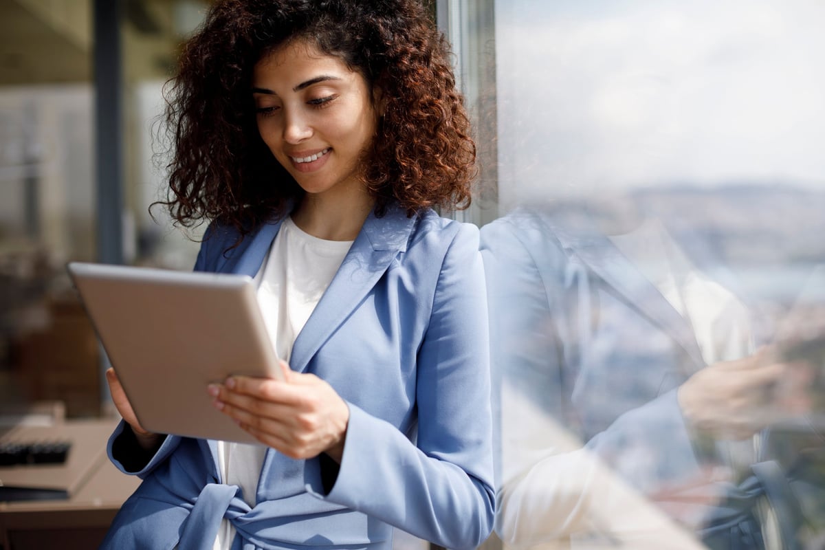 A young businesswoman leans against a window while looking at her tablet.