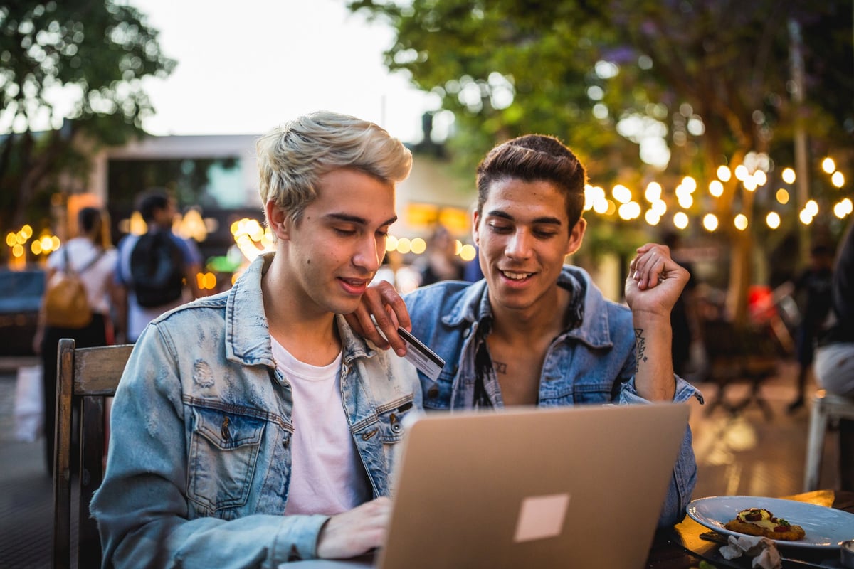 A young couple sit outside, looking at a computer. One is holding a credit card.