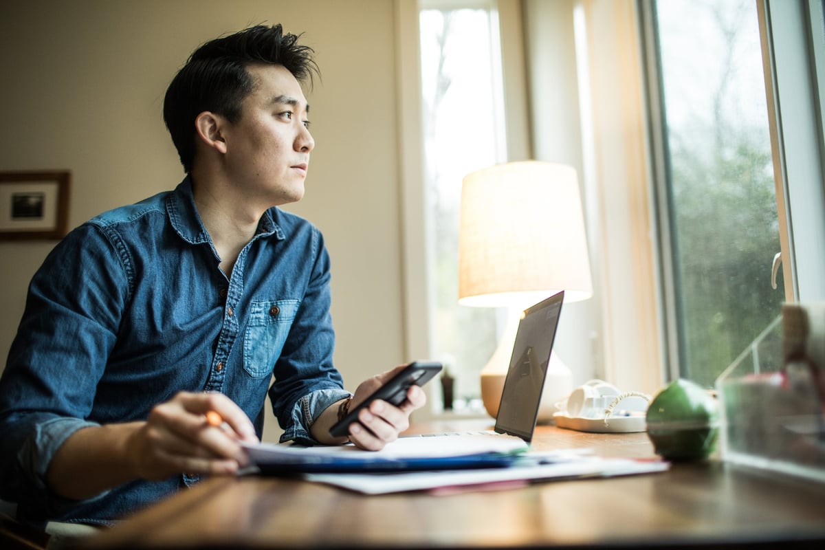 A young man sits in his home office. He is staring outside, lost in thought.