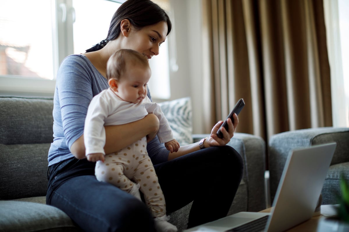 A young mother sits on the couch, working from home and holding her baby.
