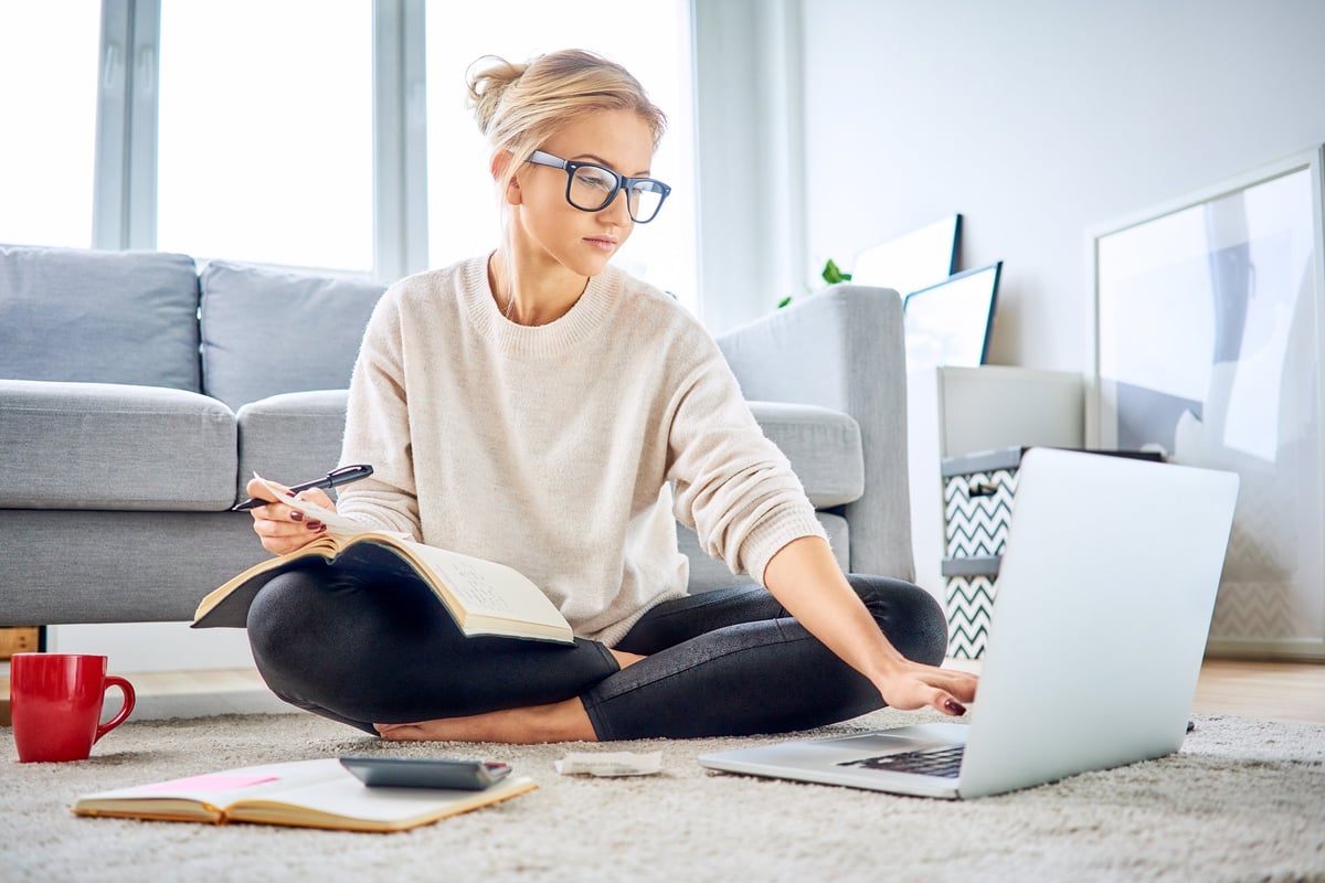A young woman sitting on the floor with a calculator, laptop, and notebook.