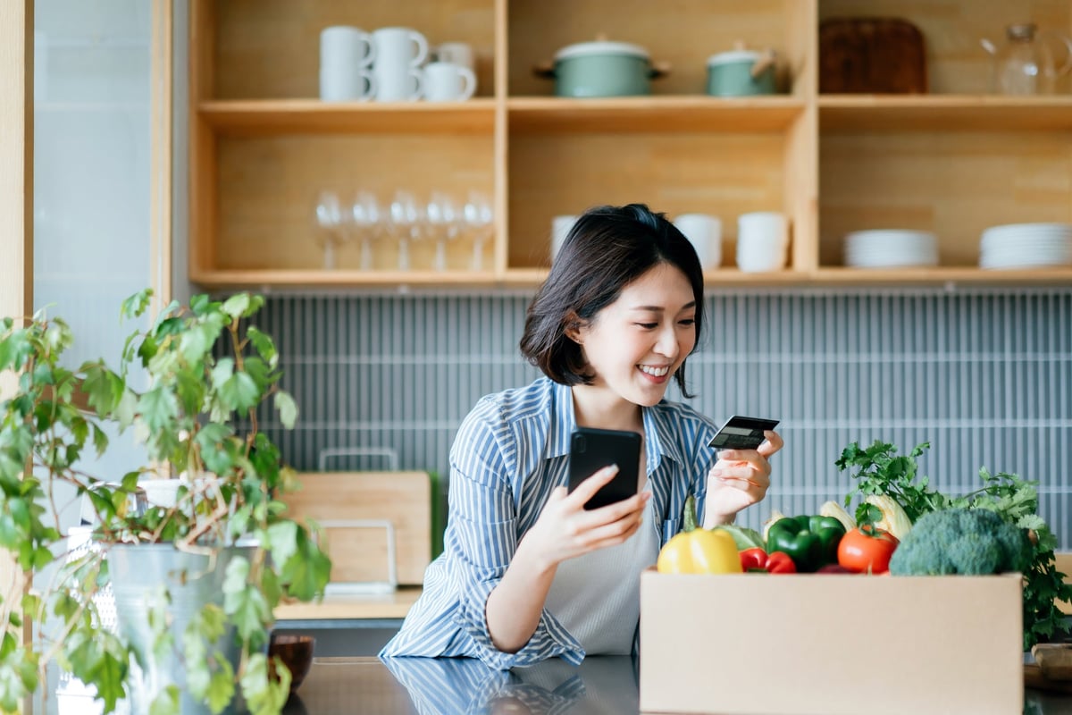 A young woman stands in her kitchen, holding her credit card and phone.