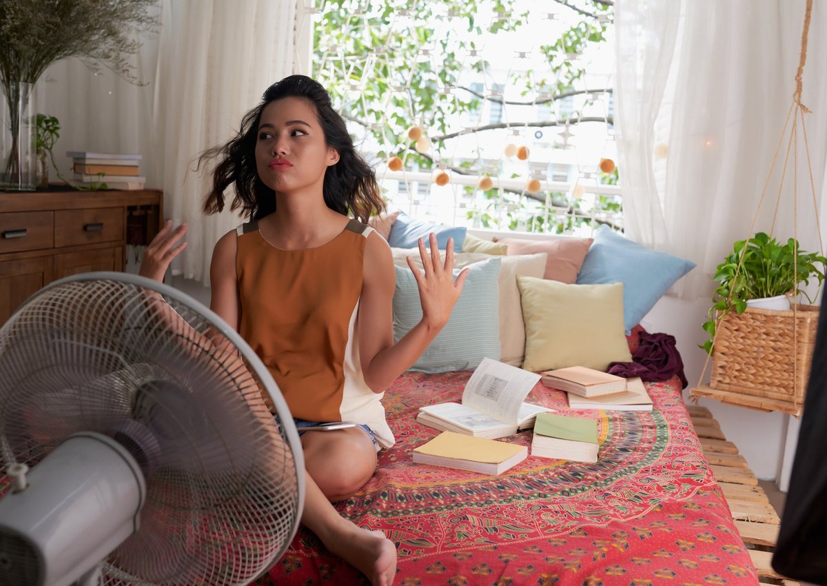A young woman tries to stay cool while sitting on her bed in front of a fan.