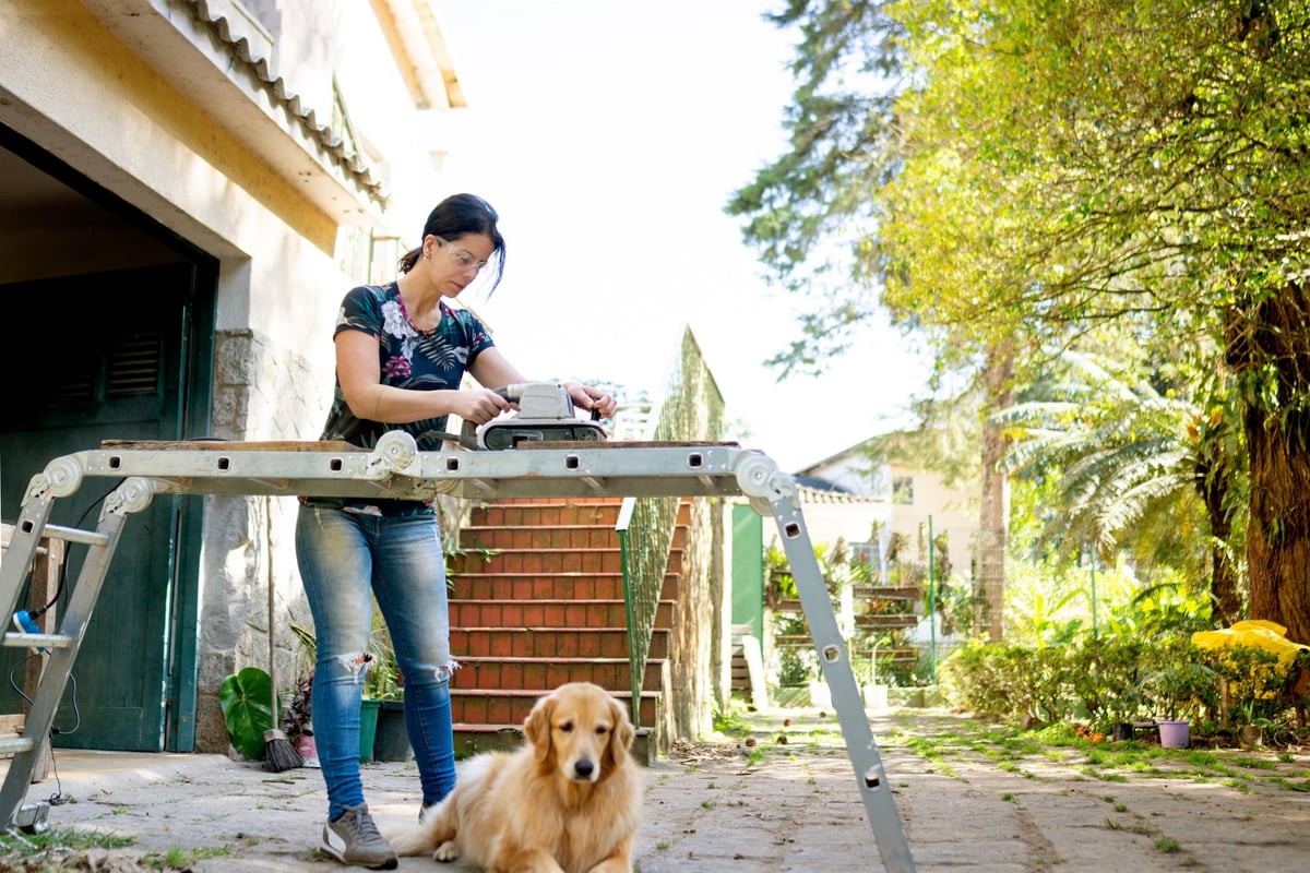 A young woman works outdoors on a home renovation project while her dog sits nearby.