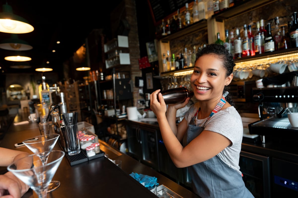 A smiling bartender shaking a cocktail shaker while standing behind a bar.