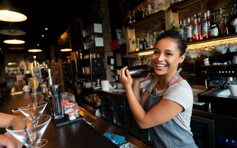 A smiling bartender shaking a cocktail shaker while standing behind a bar.