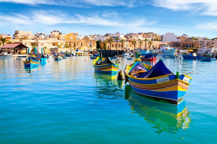Colorful boats in a harbor with bright blue water with the Malta skyline in the background.