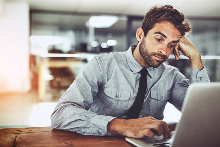 A man looking bored with his head resting on his hand while working on his computer in an office.