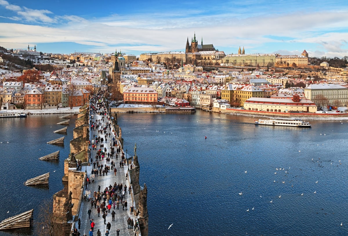 People walking across a bridge in Prague with the city and castle on the hill in the background.