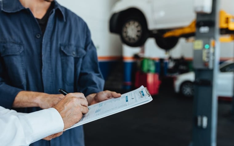 A car mechanic in a garage holds a clipboard for a customer to sign a bill for payment.