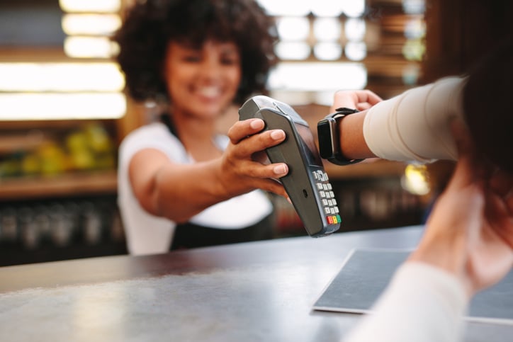 A smiling cashier holding up a payment reader for a woman paying with her smart watch.