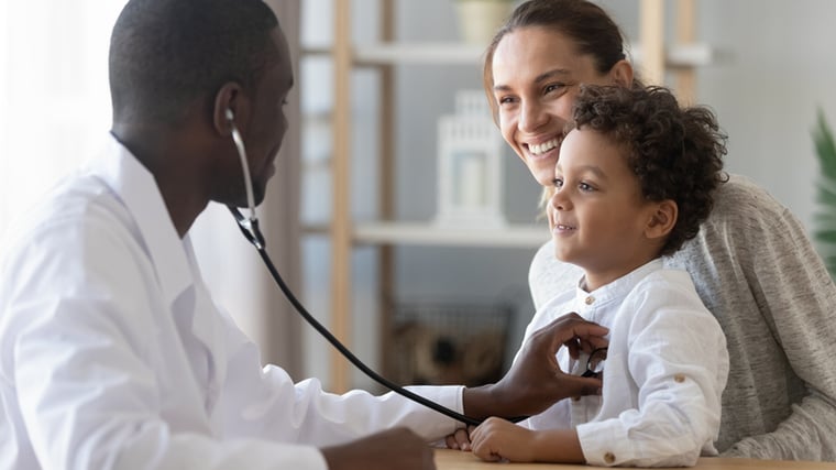 A child sitting on his mother's lap while a doctor checks his heartbeat with a stethoscope.