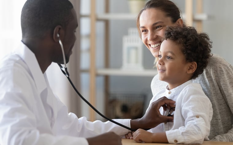 A child sitting on his mother's lap while a doctor checks his heartbeat with a stethoscope.