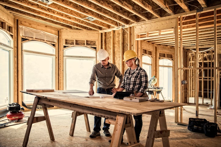 Two construction workers looking at plans on a table in a house being built.