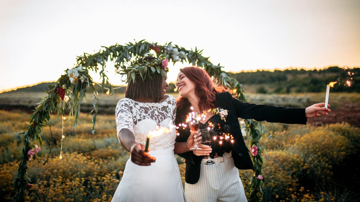 A smiling couple holding sparklers and standing in front of a flower arch at their outdoor wedding ceremony.