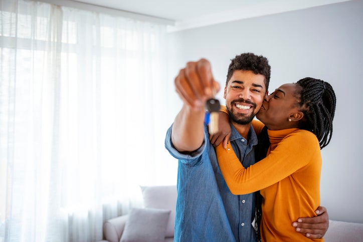 A woman kissing a smiling man's cheek while he holds up keys to the new house they're standing in.